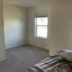 an empty bedroom with beige carpet, a bed, and two windows