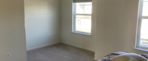 an empty bedroom with beige carpet, a bed, and two windows