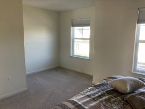 an empty bedroom with beige carpet, a bed, and two windows