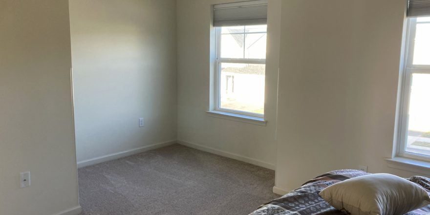 an empty bedroom with beige carpet, a bed, and two windows