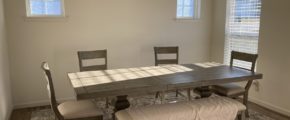 a dining room with a table, chairs, and bench in it. Bright sunlight enters through three visible windows.