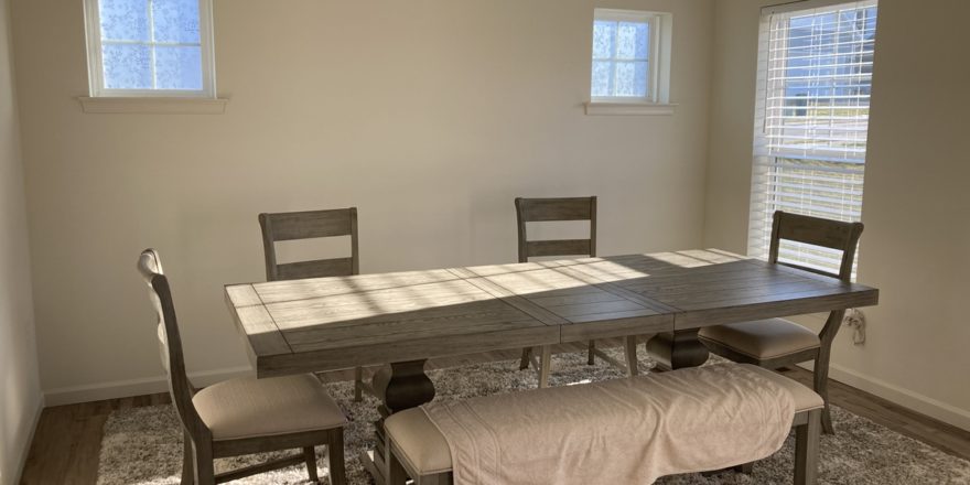 a dining room with a table, chairs, and bench in it. Bright sunlight enters through three visible windows.