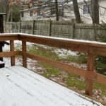 a wooden deck covered in snow with grass below and a wooden fence