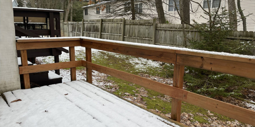 a wooden deck covered in snow with grass below and a wooden fence