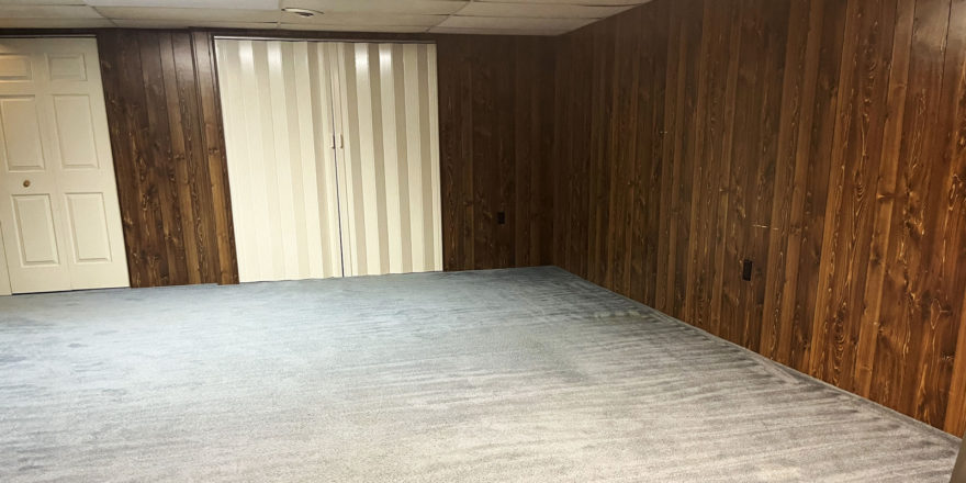 an empty room with wood paneling. beige carpet, a closet with off-white accordion doors and a second closet with off-white bi-fold doors