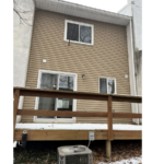 the rear of a building with a wood deck, tan siding, two white windows, and a white sliding glass door. An air conditioning unit sits below the deck.