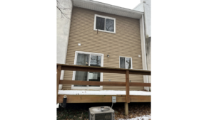 the rear of a building with a wood deck, tan siding, two white windows, and a white sliding glass door. An air conditioning unit sits below the deck.