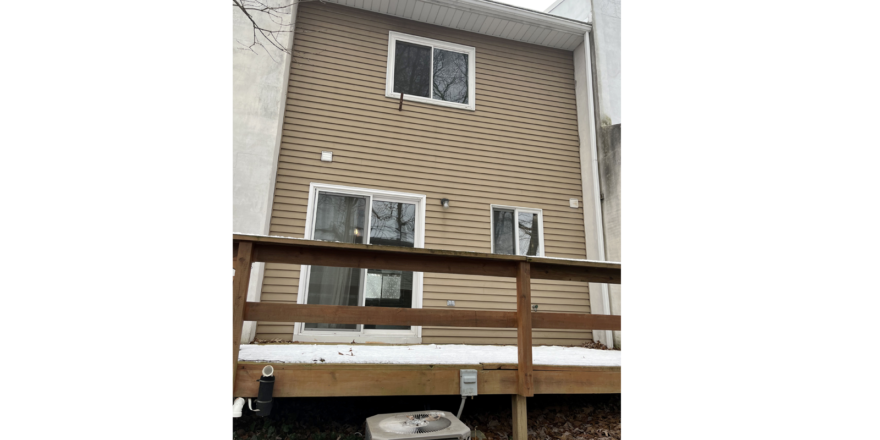 the rear of a building with a wood deck, tan siding, two white windows, and a white sliding glass door. An air conditioning unit sits below the deck.