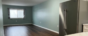 an empty living room with hard wood floors with stainless steel refrigerator in foreground