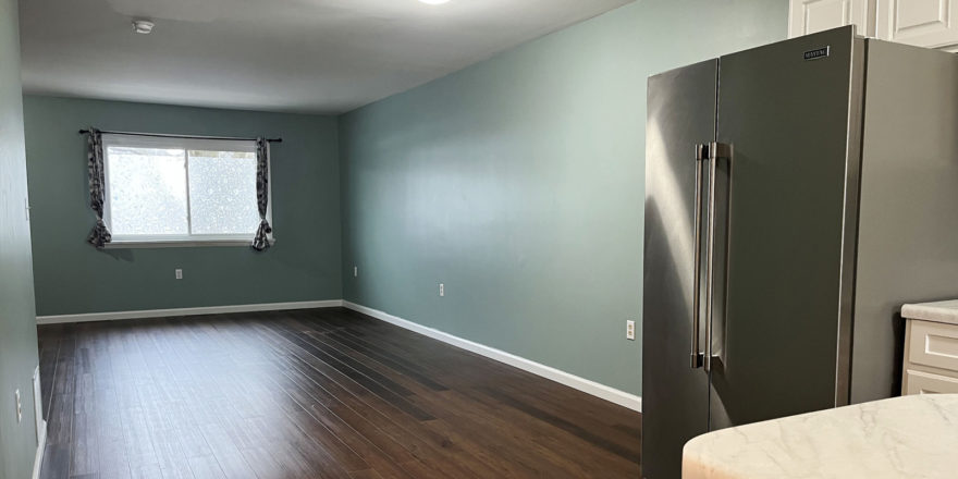 an empty living room with hard wood floors with stainless steel refrigerator in foreground