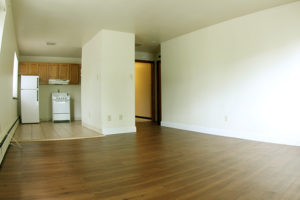 Large living area with dark wood plank flooring, off-white walls, and white baseboard trim. Entry door on right with woodgrain trim. On left, there is a kitchen with beige tile floor, white refrigerator, and white small range to the right of the refrigerator