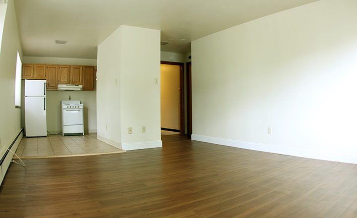 Large living area with dark wood plank flooring, off-white walls, and white baseboard trim. Entry door on right with woodgrain trim. On left, there is a kitchen with beige tile floor, white refrigerator, and white small range to the right of the refrigerator