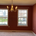 Dining room is painted a wine red and has two windows, carpet, and a chandelier