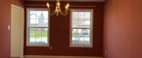 Dining room is painted a wine red and has two windows, carpet, and a chandelier