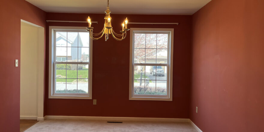 Dining room is painted a wine red and has two windows, carpet, and a chandelier