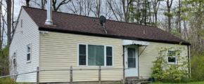 a small crean colored house with white trim, a brown shingled roof, and steel chimney