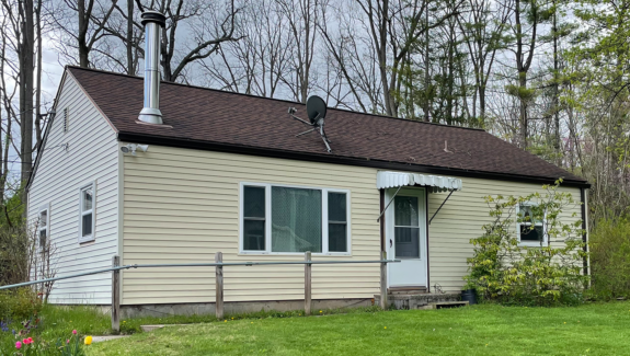 a small crean colored house with white trim, a brown shingled roof, and steel chimney