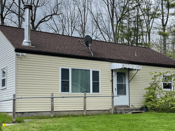 a small crean colored house with white trim, a brown shingled roof, and steel chimney