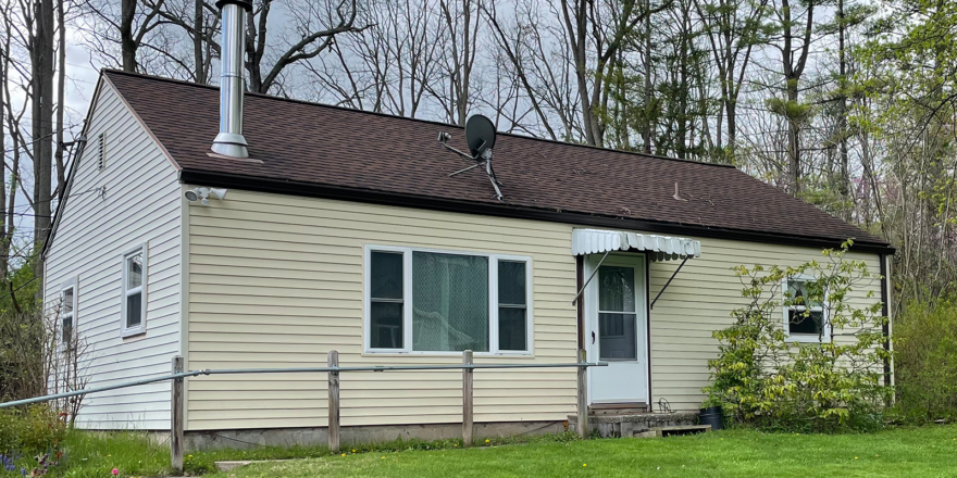 a small crean colored house with white trim, a brown shingled roof, and steel chimney